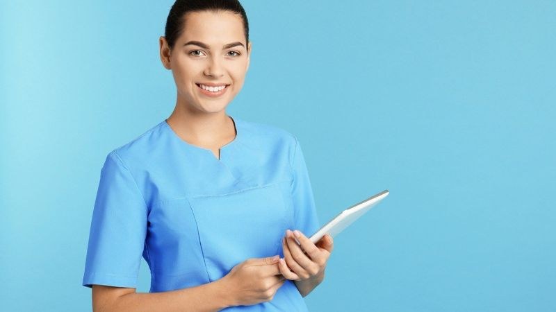 Nursing student in blue scrubs holding a tablet against a light blue background at a nursing school in San Diego, California.