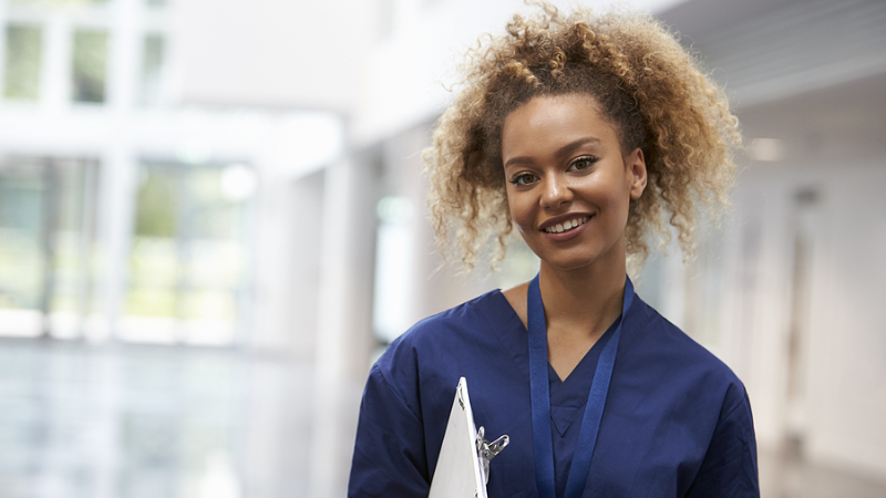Smiling nurse in blue scrubs holding a folder in a modern healthcare setting, representing students in occupational nurse practitioner programs.