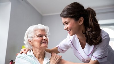 A nursing assistant in uniform smiles and speaks with an elderly woman in a home care setting, representing CNA programs in Nashville.
