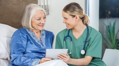 Nurse in green scrubs holding a tablet and smiling at an older woman sitting in bed, illustrating the supportive role taught in CNA programs in Austin, Texas.